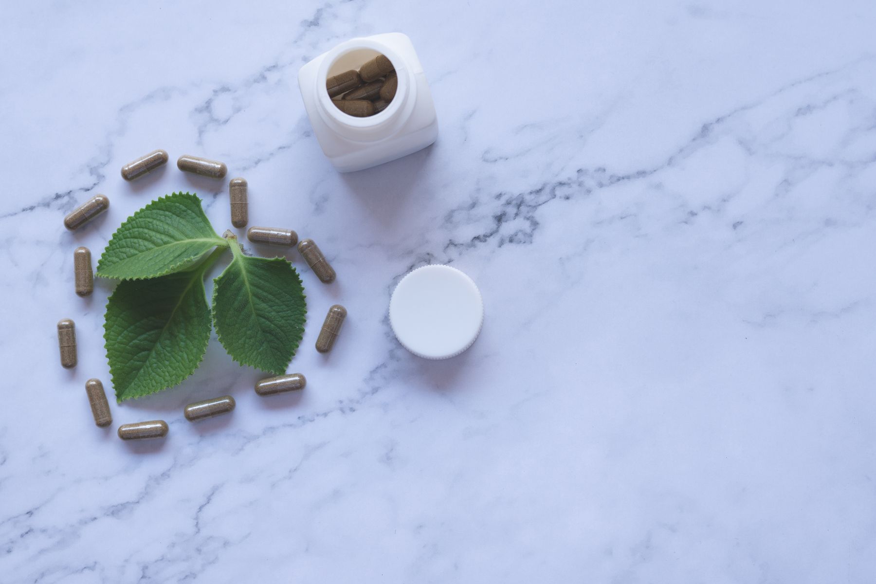 Herbal capsules arranged around fresh green leaves beside an open bottle